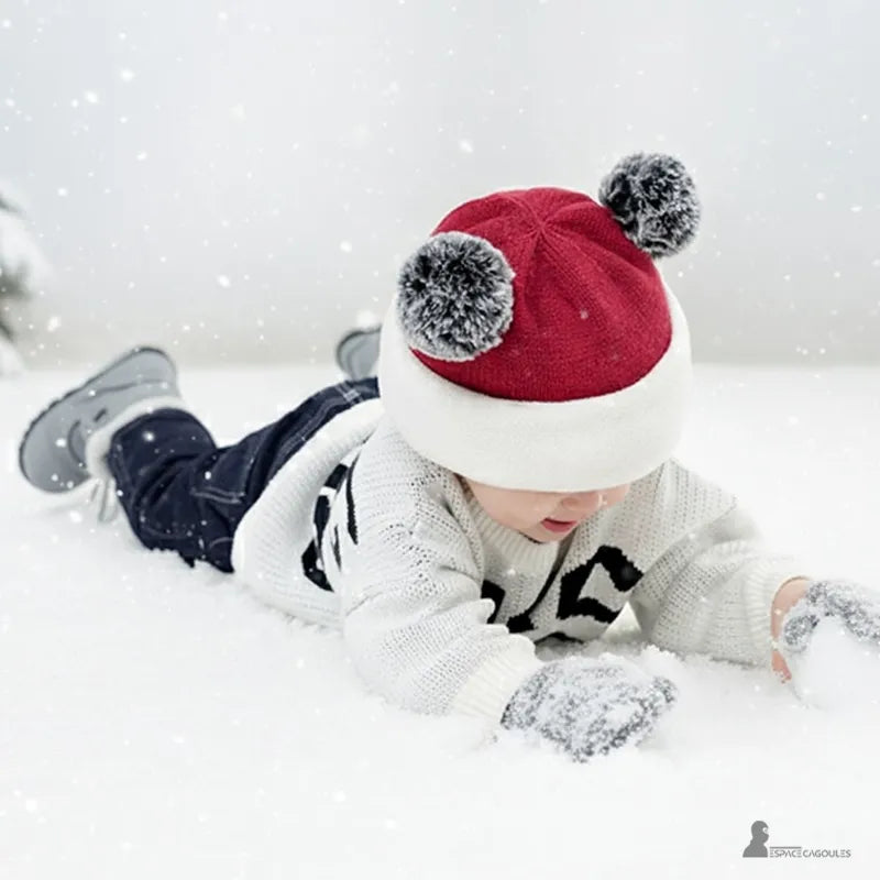 Cagoule tricot enfant rouge et blanche avec pompons, petit garçon joue dans la neige en plein air couché sur le sol