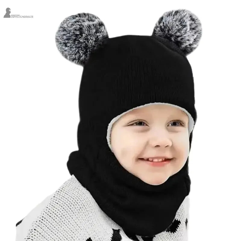 Cagoule tricot enfant noire avec pompons gris, petit garçon souriant en intérieur portant un pull blanc à motifs noirs