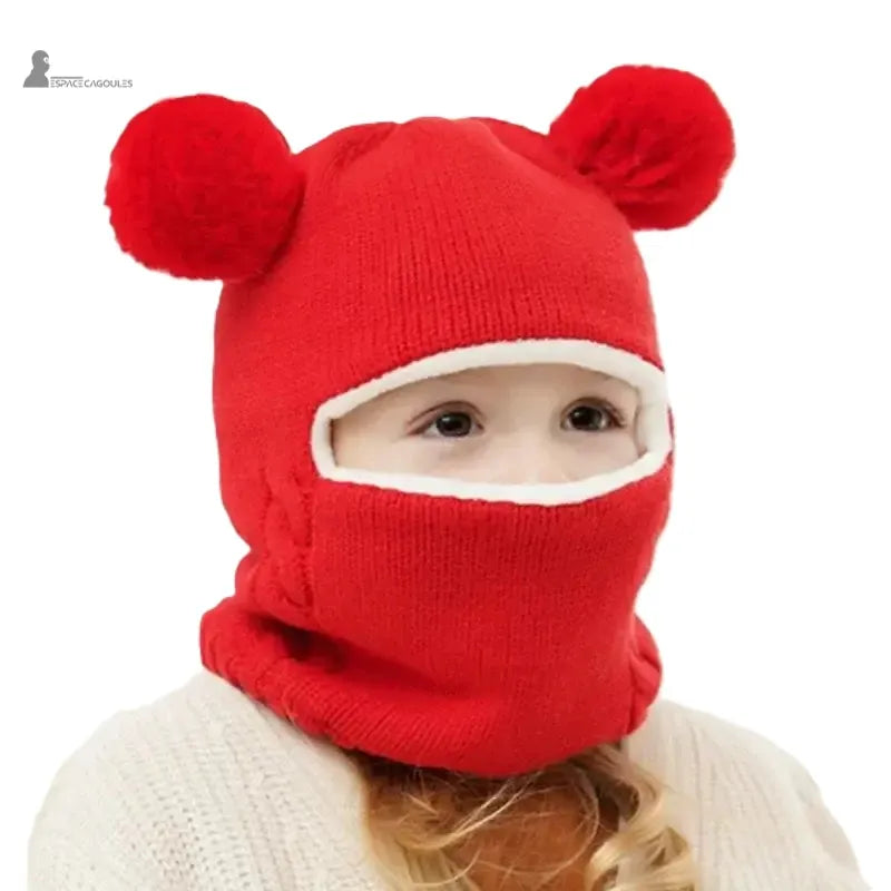 Cagoule tricot enfant rouge avec deux pompons ronds, fillette portant la cagoule relevée sur le visage en intérieur
