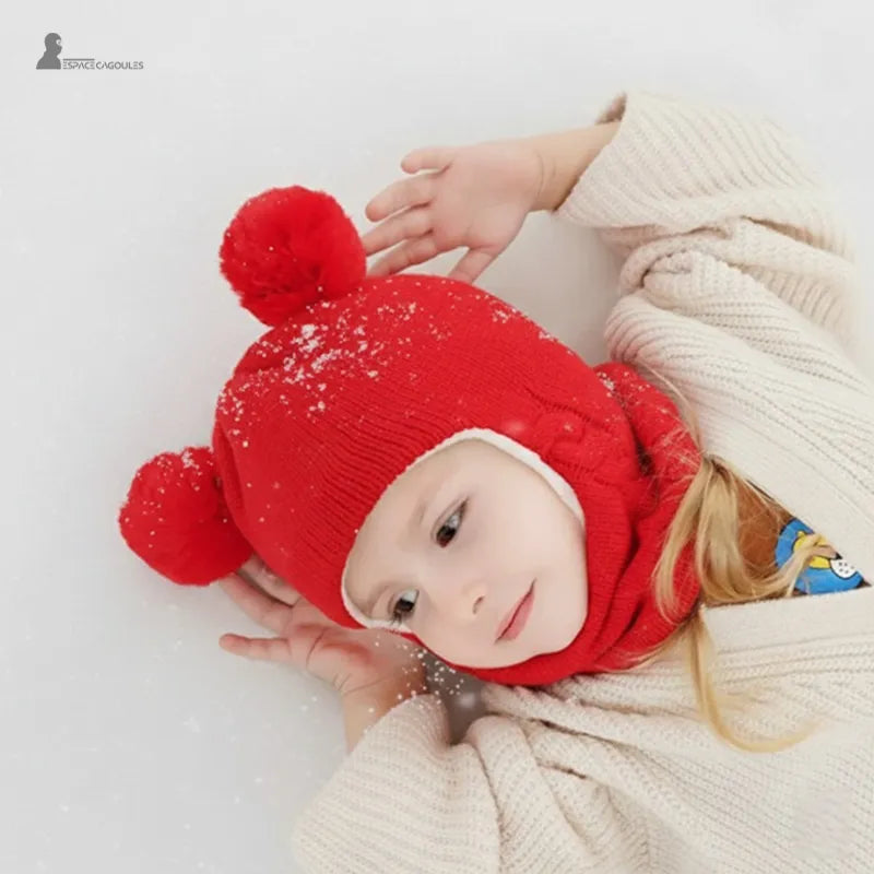 Cagoule tricot enfant rouge avec pompons, fillette allongée dans la neige jouant dehors avec flocons sur la cagoule