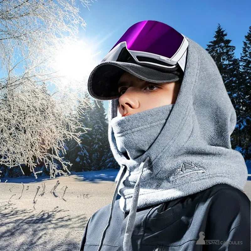 Grey women's balaclava hood, worn outdoors, with a mountain logo, under the snow. A person is in profile, wearing ski goggles.