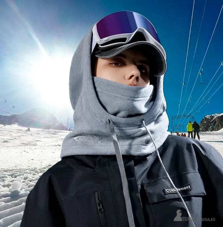 Grey women's balaclava hood, worn outdoors in the mountains, in fleece fabric with a logo. A person is in front, wearing ski goggles, a black jacket and a cap.