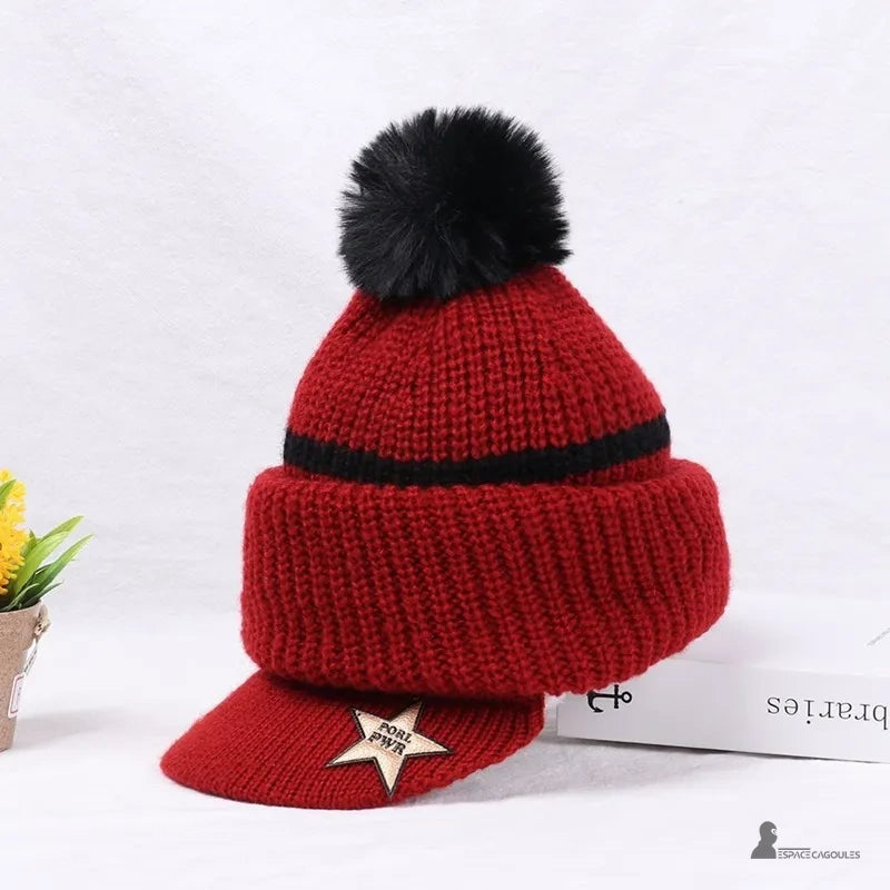 Casquette bonnet enfant rouge en laine avec pompon noir et visière, posé sur fond blanc avec une boîte et une fleur décorative