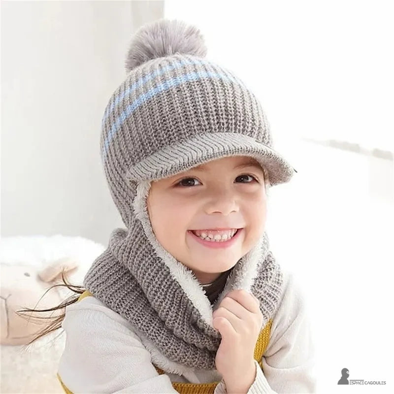 Casquette bonnet enfant grise en laine avec pompon et rayures bleues, petite fille souriante dans une chambre claire