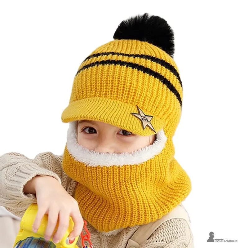 Casquette bonnet enfant jaune tricot avec pompon noir et rayures noires, petit garçon joue avec un jouet coloré en intérieur