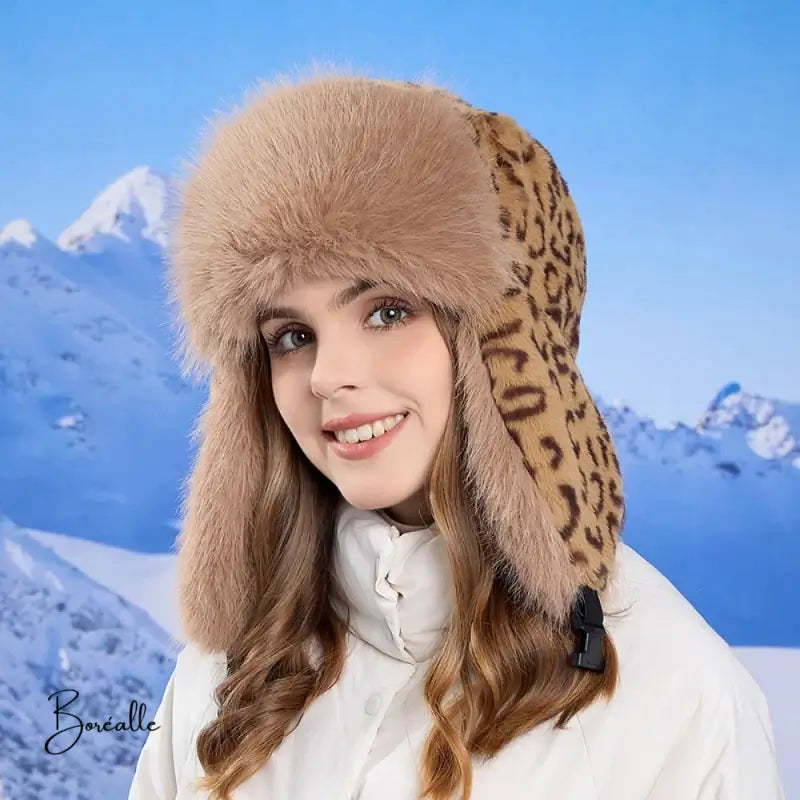 Lächelnde Frau, die eine kaffeebraune Leopardenfrau, einen russischen Hut mit Leoparden-Kunstpelz-Innenseite und Ohrenklappen trägt. Sie ist warm angezogen und posiert vor einer Kulisse aus schneebedeckten Bergen.