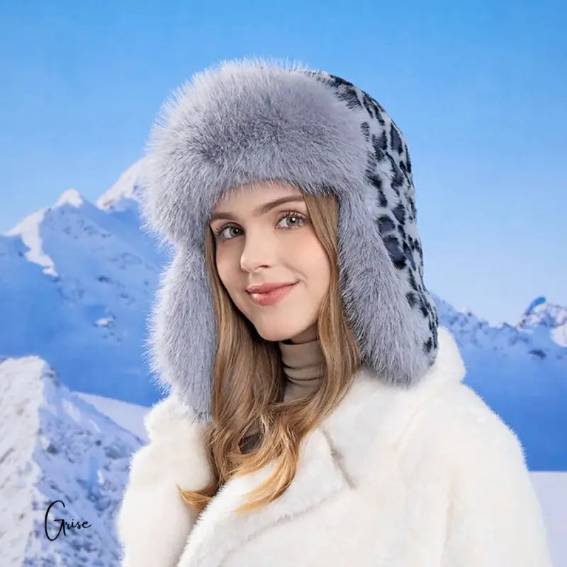 Lächelnde Frau trägt einen grauen russischen Damenhut mit Leopardenmuster und Ohrklappen. Hintergrund schneebedeckter Berge, Winteratmosphäre.
