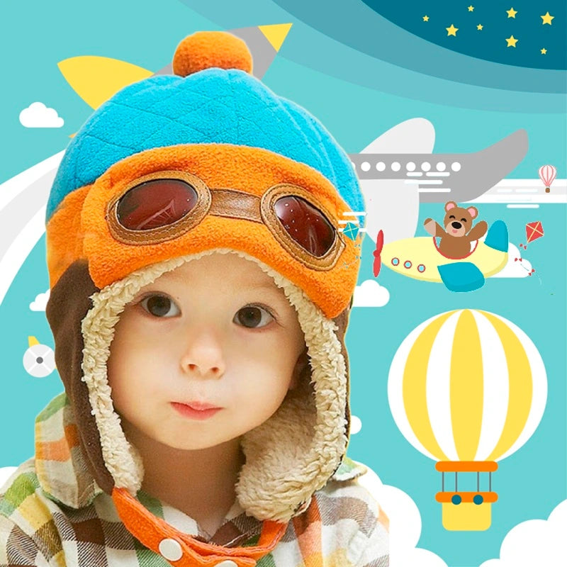 Child with a blue and orange aviator hat, warm lining and glasses, against a background of aerial scenery with hot air balloon.