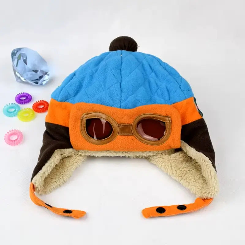 Blue, orange and brown aviator children's hat with aviator glasses, placed on a white background and decorated with colorful buttons.
