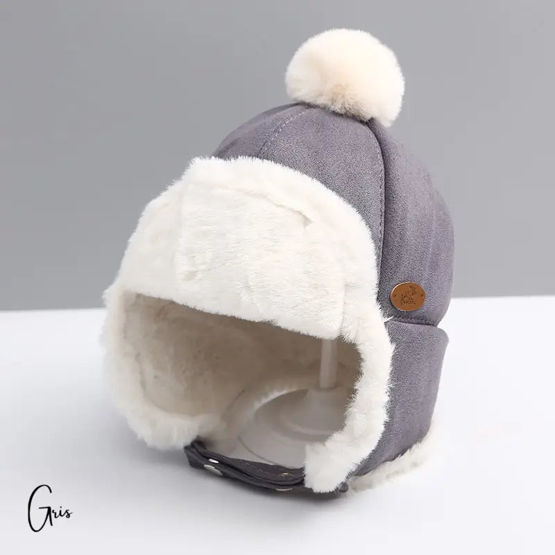 Children's chapka in grey suede with white pompom and soft faux fur interior, seen from the back on a neutral background. Ideal winter protection with an aviator design.