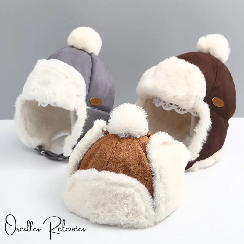 Three children's chapkas in brown and grey suede with faux fur interior and fluffy pompom, set on a light surface. Warm and cozy model for winter.