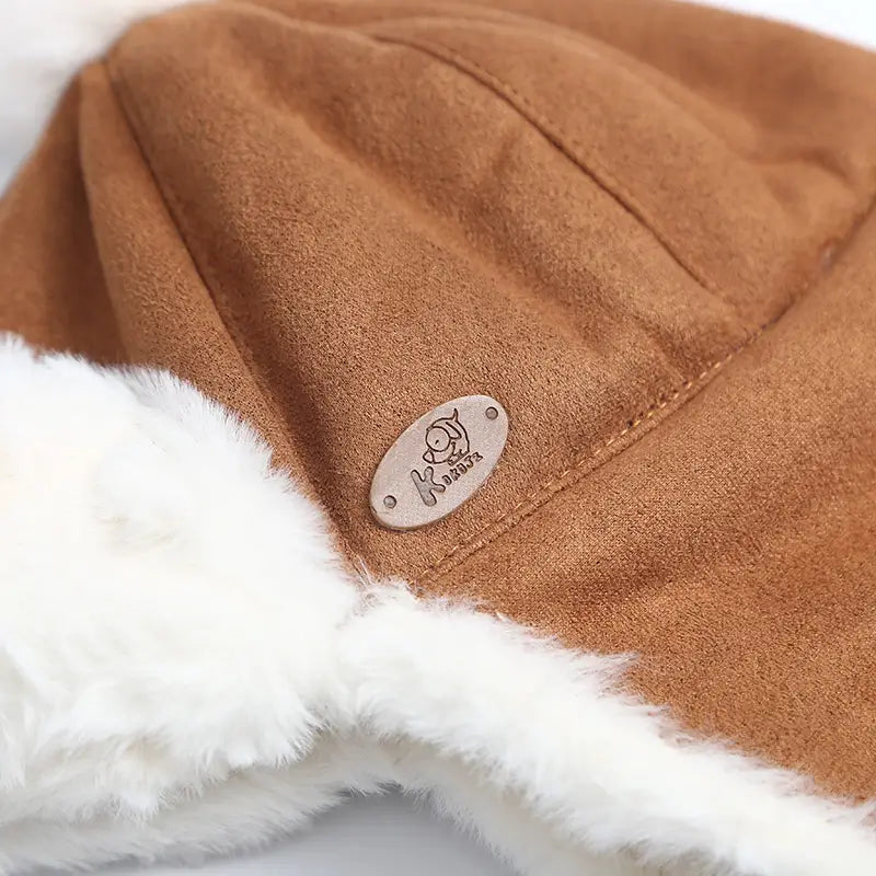 Zoom on the brown suede texture of a Children's Chapka with white faux fur interior and a small metal patch. Elegant and soft finishes for winter.