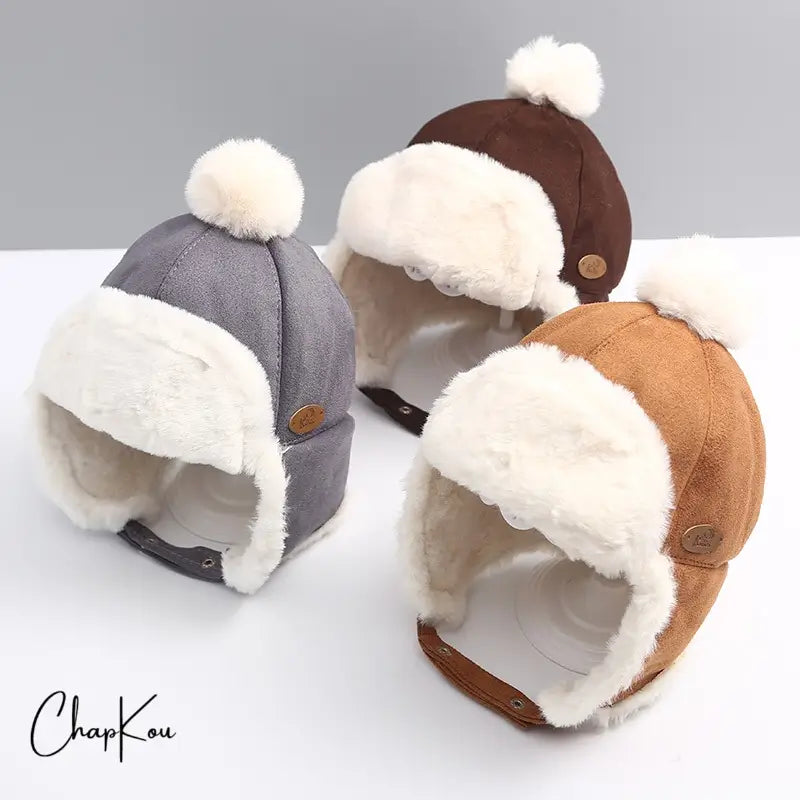 Three brown and grey suede children's chapkas with white faux fur interior and pompom, arranged on a light surface. Ideal for protecting from the cold with a retro aviator design.
