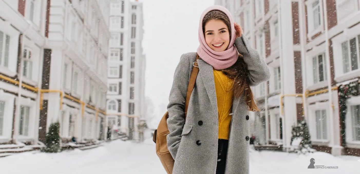 Cagoule femme rose pâle en laine douce portée avec un manteau gris et un pull jaune, femme souriante marchant dans une rue enneigée – Cagoule Femme - Espace Cagoules