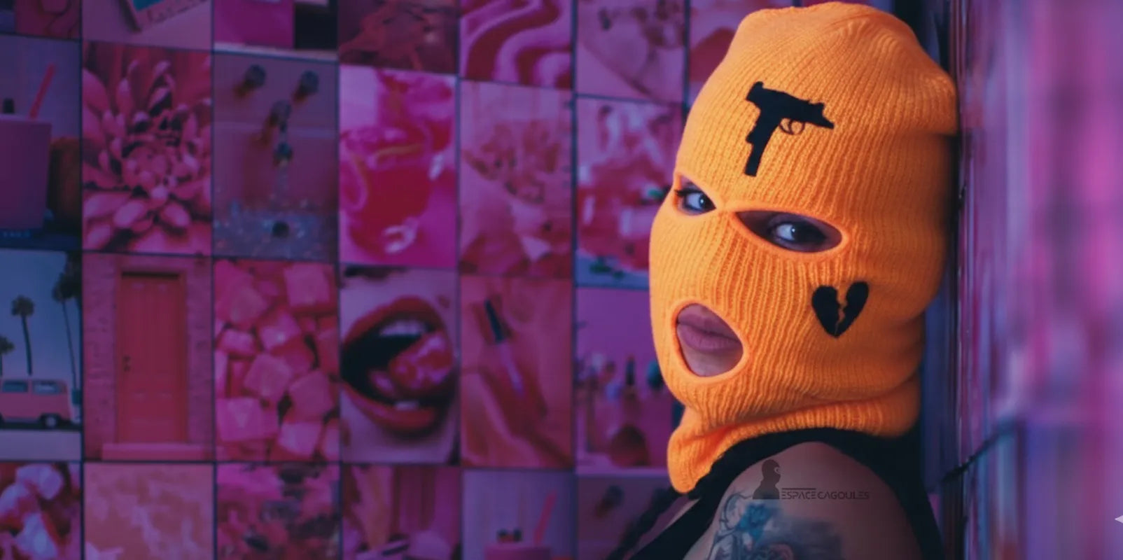 Cagoule fluo orange avec motifs noirs en forme de cœur et pistolet, femme posant devant un mur rose vif, ambiance artistique et colorée