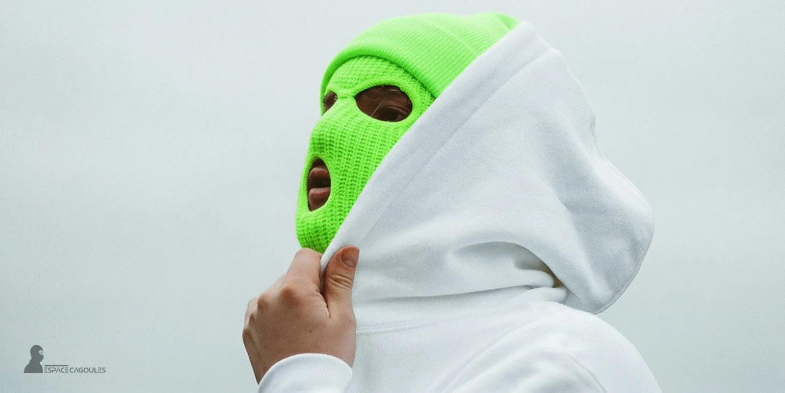 Cagoule fluo verte sur fond clair portée par un rappeur avec un sweat à capuche blanc, visage partiellement couvert, atmosphère urbaine minimaliste
