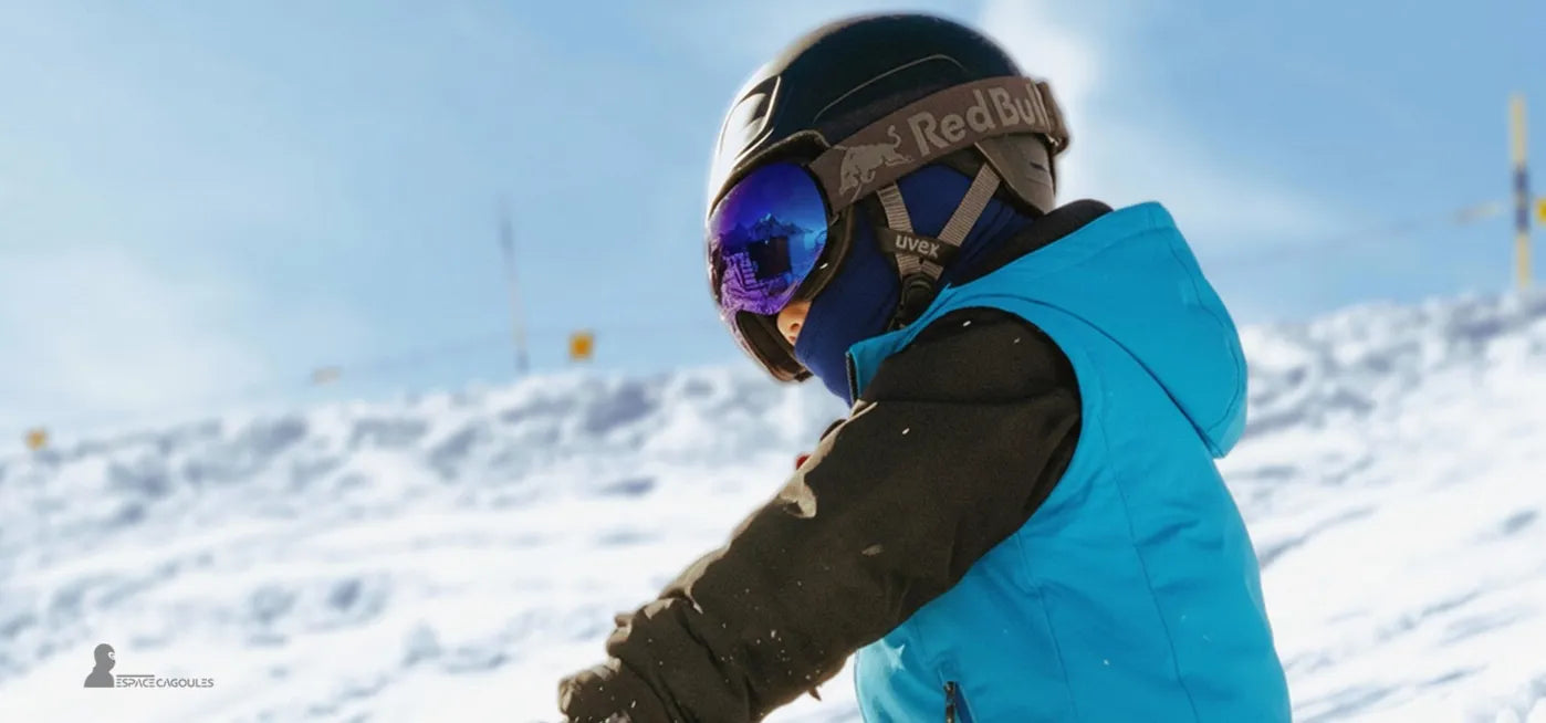 Cagoule ski bleue ajustée sous un casque noir portée par un enfant en veste turquoise jouant sur une piste enneigée par ciel bleu – Espace Cagoules