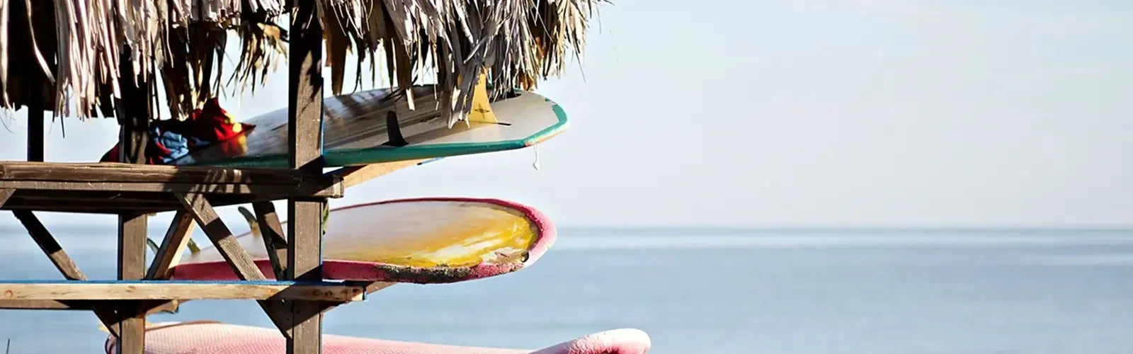 Planches de surf rangĂ©es sous une paillote face Ă  la mer, illustrant la collection Cagoules de Surf â Espace Cagoules