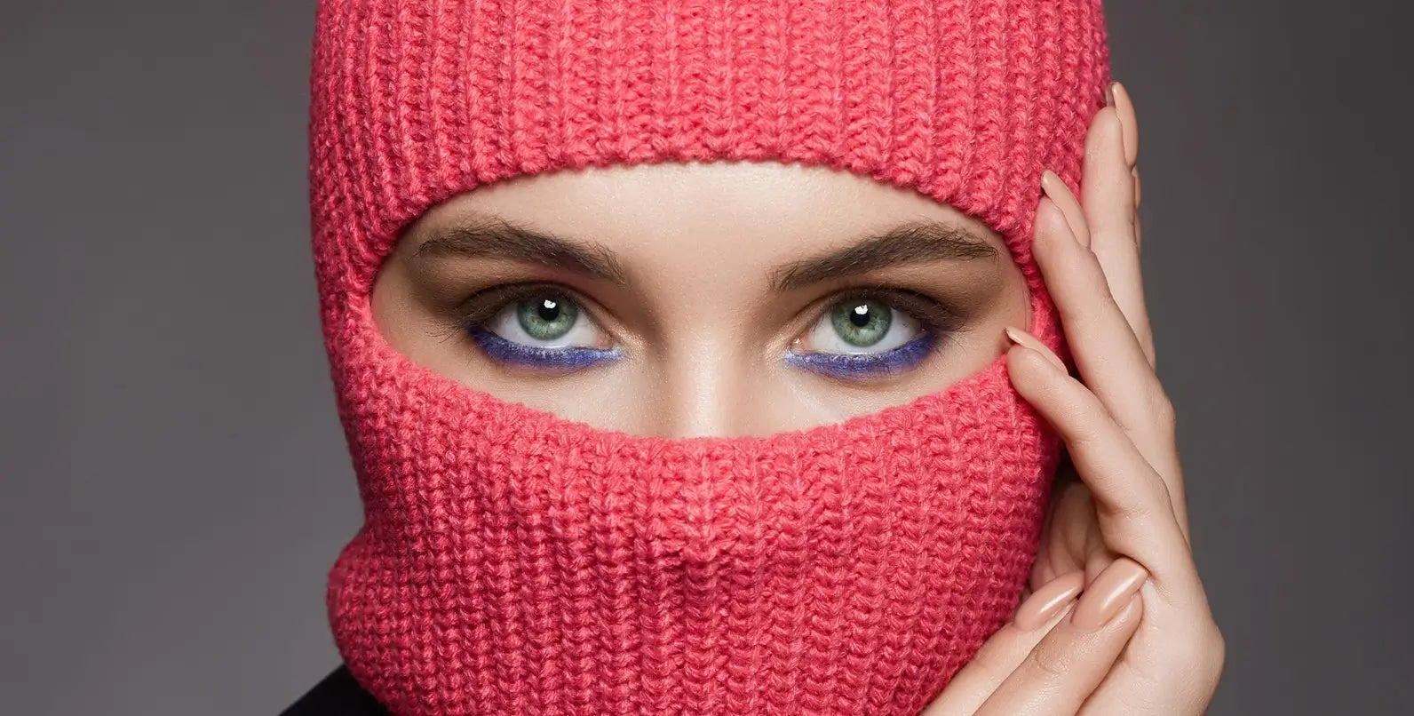 Gros plan sur une femme portant une cagoule rose en maille, mettant en valeur ses yeux verts maquillés d'un trait violet, avec une pose élégante et contemporaine