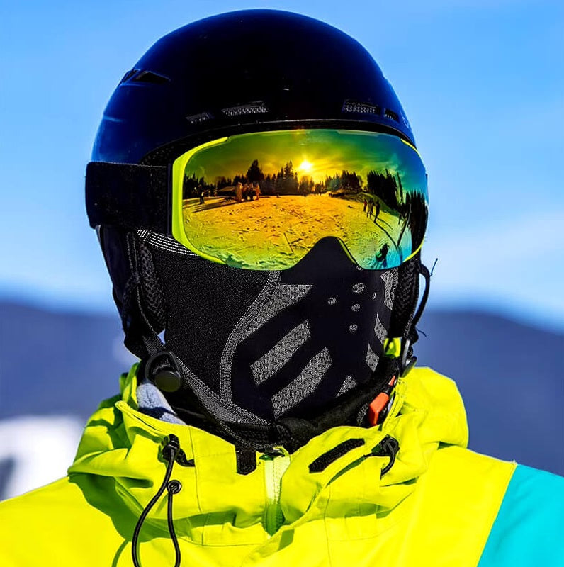 Homme portant une cagoule pour le ski noire avec des motifs réfléchissants, un casque et des lunettes de ski avec reflets de paysage enneigé au coucher de soleil, vêtu d'une veste jaune vif.