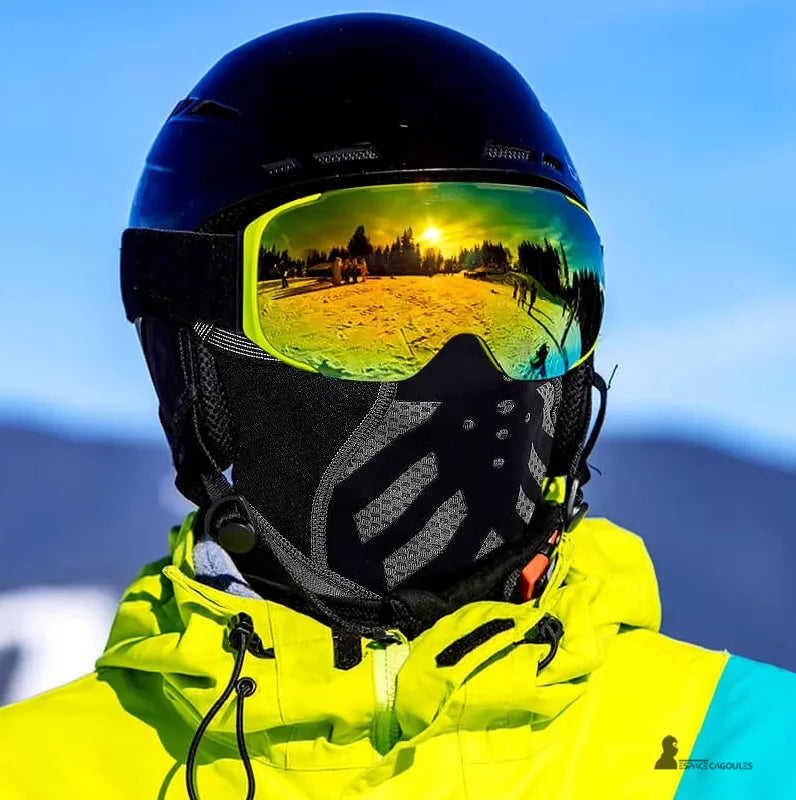 Cagoule pour le ski noire portée avec casque et masque de ski réfléchissant au coucher de soleil en montagne, offrant protection et confort.