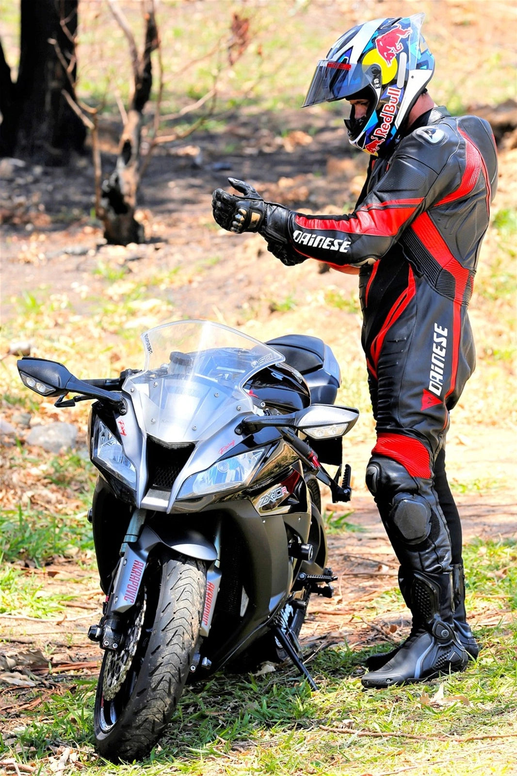 Motard en combinaison Dainese ajustant ses gants à côté d'une moto sportive noire dans un environnement naturel