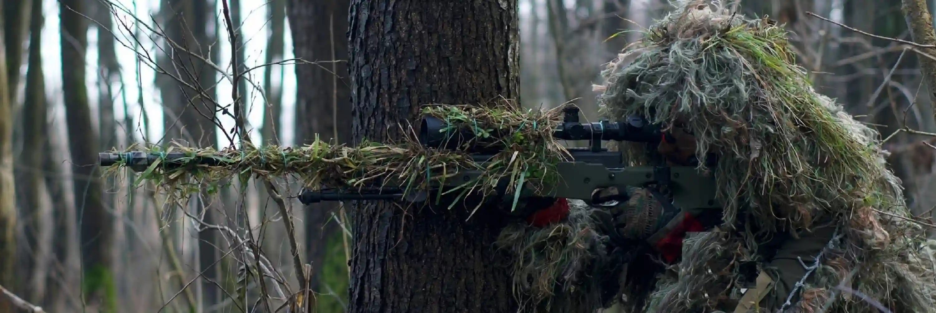 Cagoule camouflage – tireur embusqué équipé d’une tenue ghillie et d’un fusil camouflé, dissimulé dans une forêt pour illustrer la collection de cagoules camouflage.