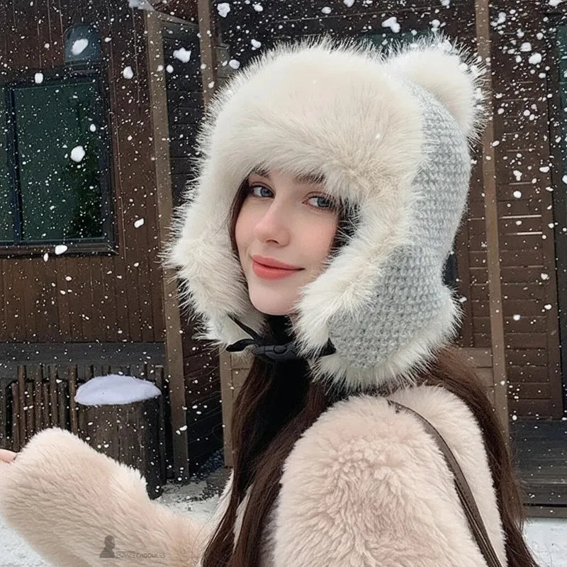 Bonnet chapka fourrure porté par une femme devant un chalet en bois sous la neige, modèle gris clair avec large contour en fausse fourrure blanche, vue de trois-quarts – Espace Cagoules