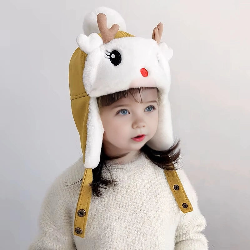Petite fille portant un bonnet enfant en polaire blanche et jaune avec des cornes marron et un pompon, fixant l'objectif avec un manteau d'hiver.