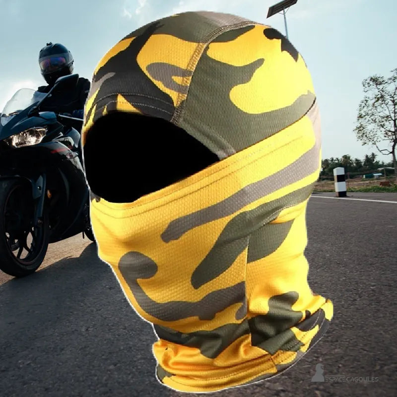 Cagoule balaclava moto camouflage jaune et vert posée sur route avec un motard et sa moto en arrière-plan