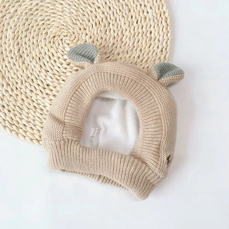 Cagoule bébé 6 à 36 mois couleur beige posée sur un tapis en osier. Design en tricot épais avec des oreilles décoratives, ouverture arrondie pour le visage et bordures côtelées assurant un bon maintien.