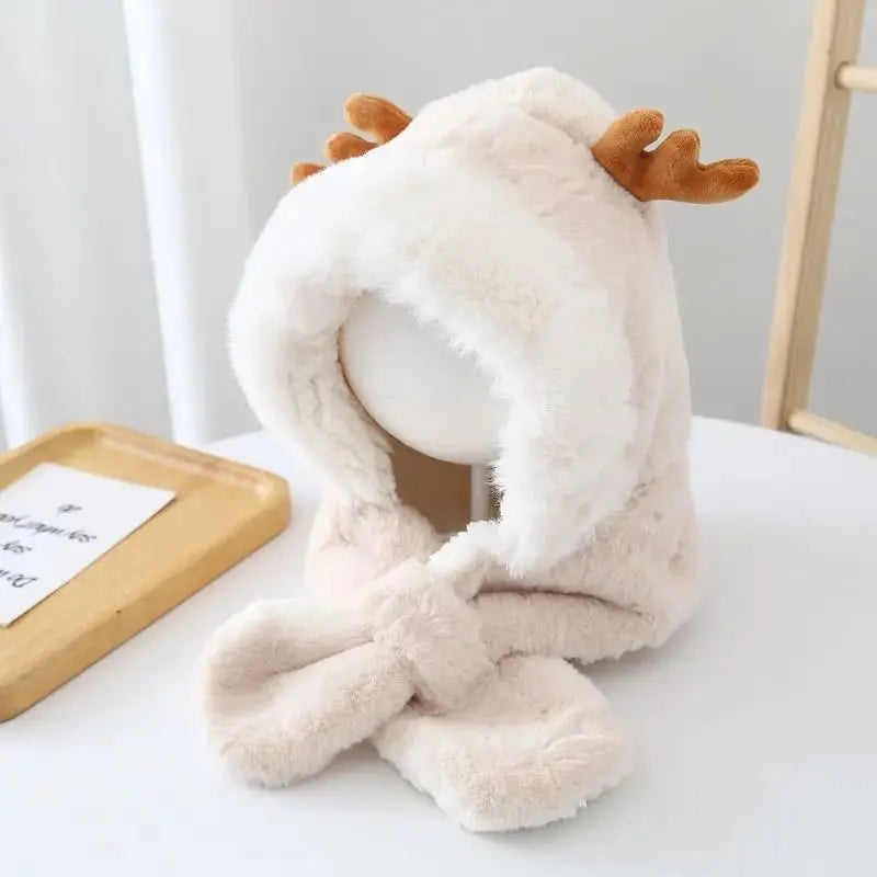 Cagoule bébé fille en fausse fourrure beige avec des cornes de cerf, posée sur un mannequin en intérieur. Écharpe intégrée croisée sous le menton, exposée à côté d’une table.