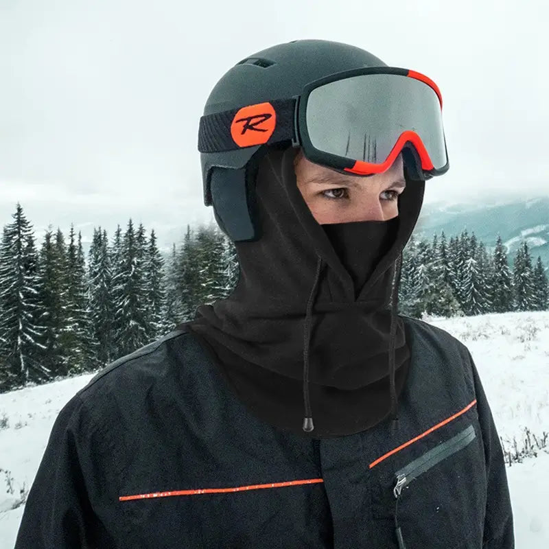 Personne équipée d'une cagoule polaire chaude noire sous un casque de ski, avec des lunettes de protection aux reflets rouges. Le design couvrant protège le visage et le cou contre le froid extrême. Paysage enneigé en arrière-plan avec des sapins sous un ciel gris.