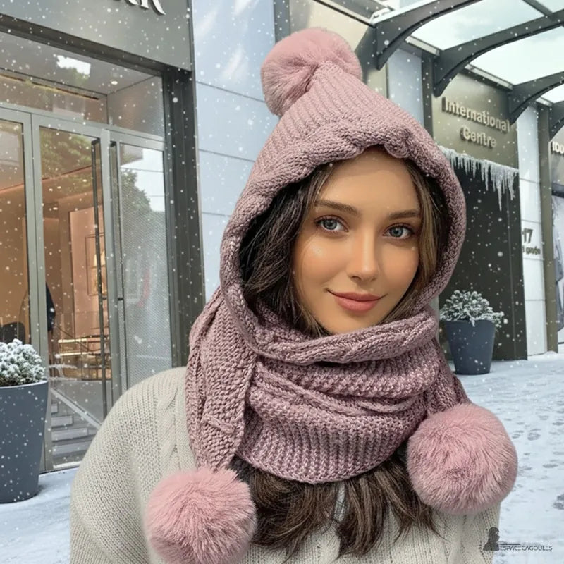 Cagoule écharpe rose poudré en laine tricotée avec pompons, femme souriante devant un centre commercial sous la neige – Espace Cagoules