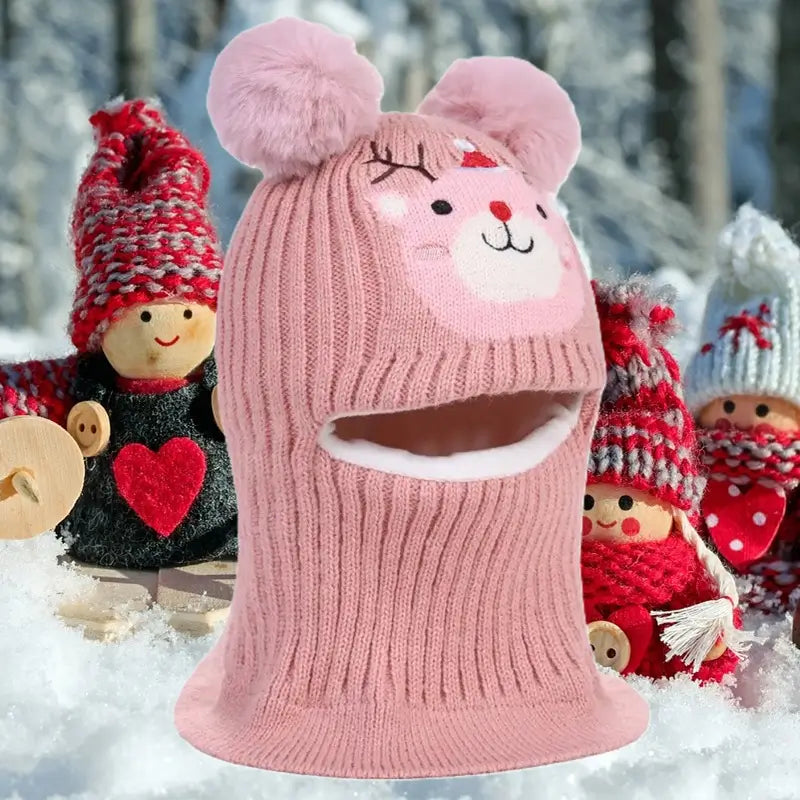 Cagoule enfant 5 ans en tricot rose avec pompons et motif animal mignon, parfaite pour l'hiver, posée dans un décor enneigé avec des poupées en arrière-plan.