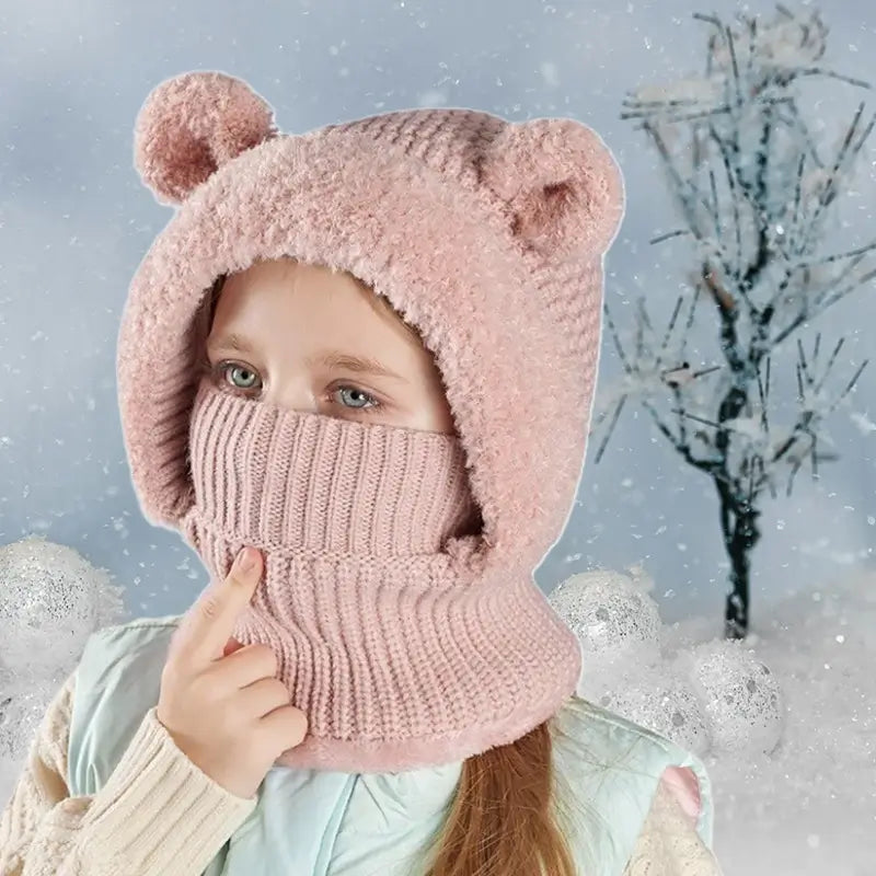 Cagoule enfant fille rose en tricot, avec des oreilles d'ours sur la capuche, couvrant le visage pour une protection optimale contre le froid, portée dans un décor hivernal enneigé.