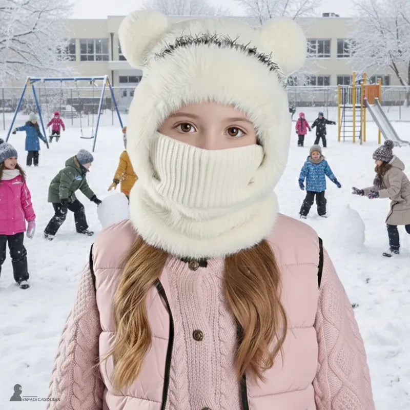 cagoule enfant fille – Enfant portant une cagoule blanche relevée avec masque tricoté blanc, en extérieur dans une cour enneigée avec autres enfants jouant – Espace Cagoules