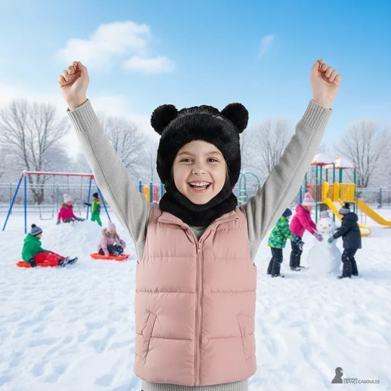 cagoule enfant fille – Fillette souriante portant une cagoule noire à oreilles d’ours, bras levés, terrain de jeu enneigé en arrière-plan avec enfants jouant dans la neige – Espace Cagoules