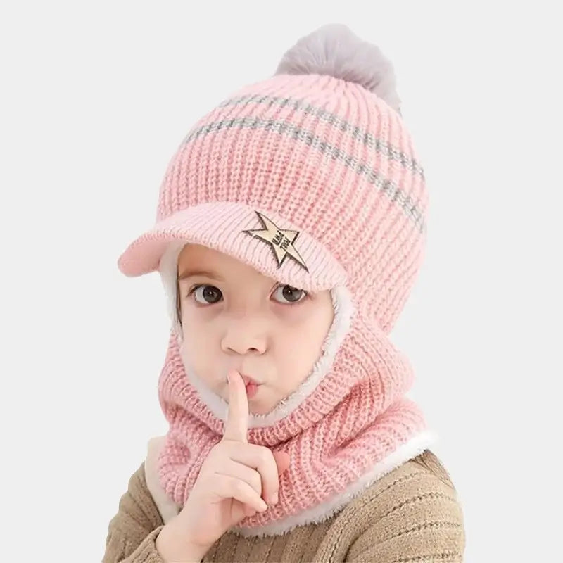 Enfant portant une cagoule enfant tricotée rose avec pompon gris, visière étoilée et doublure blanche moelleuse, posant avec un doigt sur les lèvres dans une attitude joueuse.