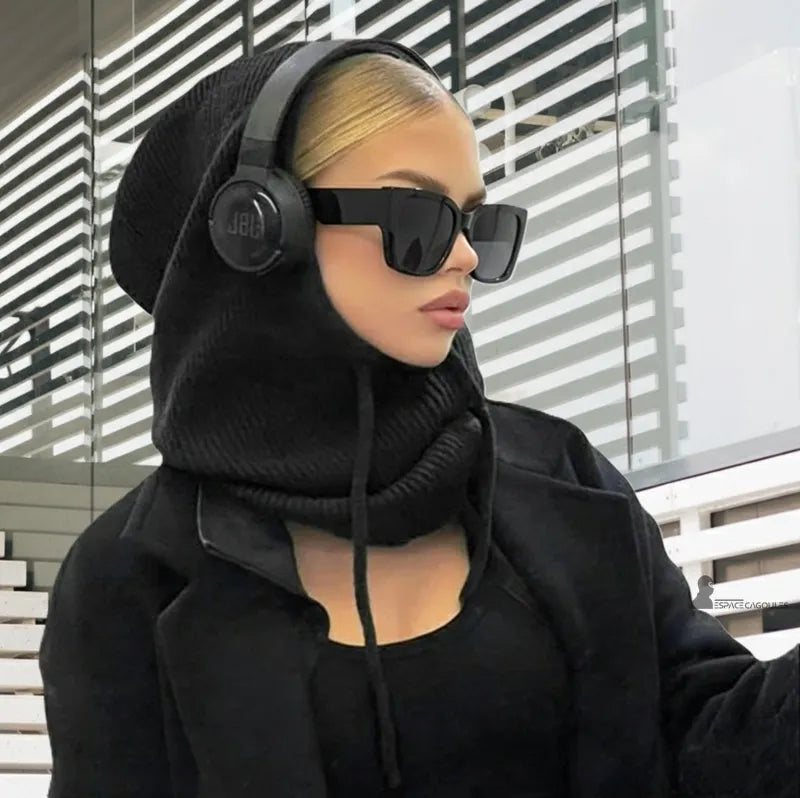 Passamontagna invernale donna nera indossata da una donna bionda con cuffie e occhiali neri, seduta in un ambiente moderno con linee architettoniche - Spazio Passamontagna.