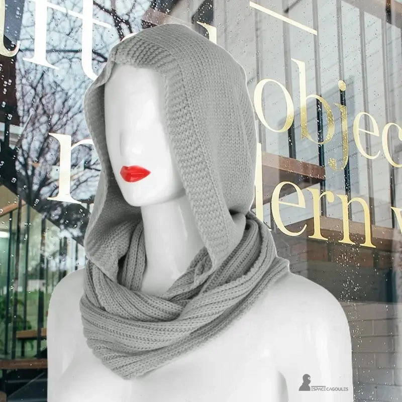Cagoule femme mode grise clair douce et chaude en maille tricotée, exposée sur mannequin devant une vitrine urbaine sous la pluie