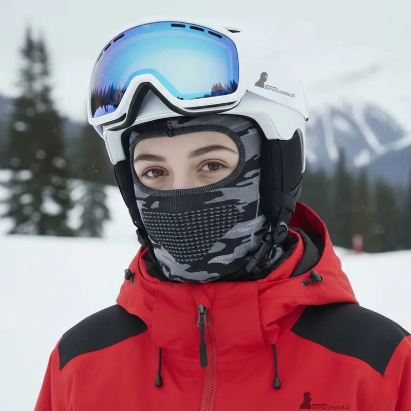 Cagoule fine sous casque de ski camouflage gris portée par une skieuse en casque blanc et masque, veste rouge, en environnement de montagne enneigé - Espace Cagoules