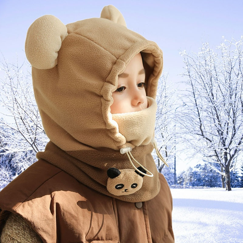 Profil d'un enfant portant une cagoule grand froid enfant beige avec oreilles d'ours, col chaud orné de pattes d'ours, en paysage hivernal enneigé.