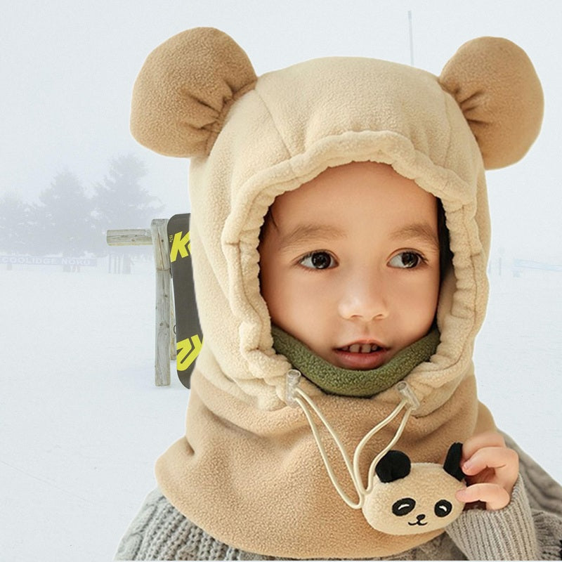 Enfant vêtu d'une cagoule grand froid enfant beige avec oreilles d'ours, sourire visible malgré le froid, fond de neige et arbres d'hiver.