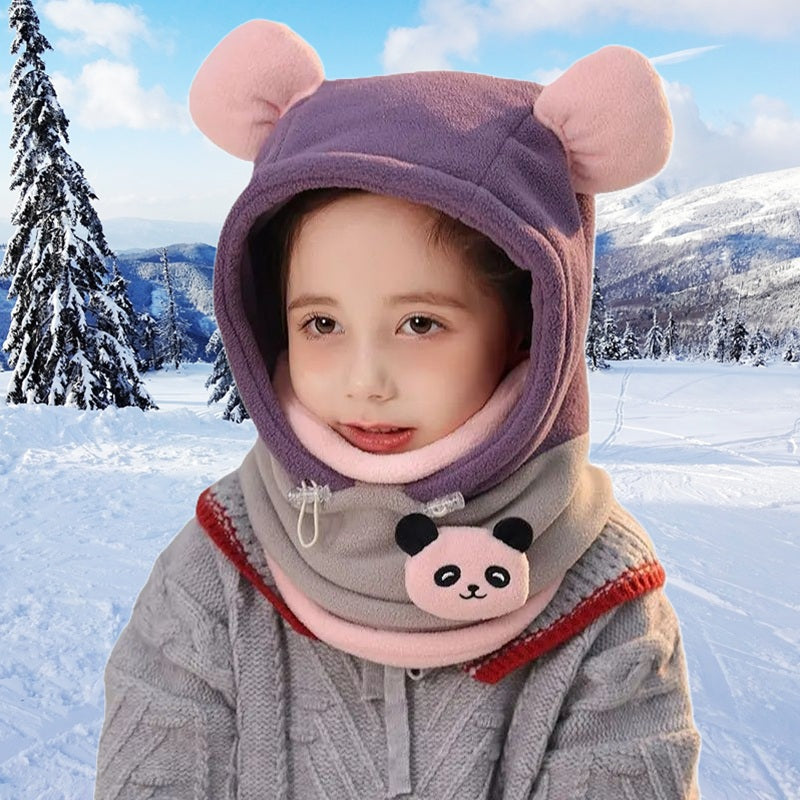 Enfant souriant portant une cagoule grand froid enfant violette et rose avec oreilles d'ourson et col orné d'un motif panda, en extérieur enneigé avec sapins et montagnes en fond.