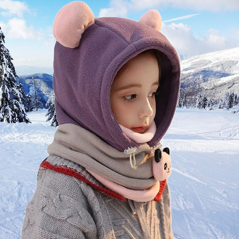 Enfant habillé d'une cagoule grand froid enfant violette et rose avec oreilles rondes, col chaud rayé et décor panda, debout sur une piste enneigée sous un ciel bleu d'hiver.