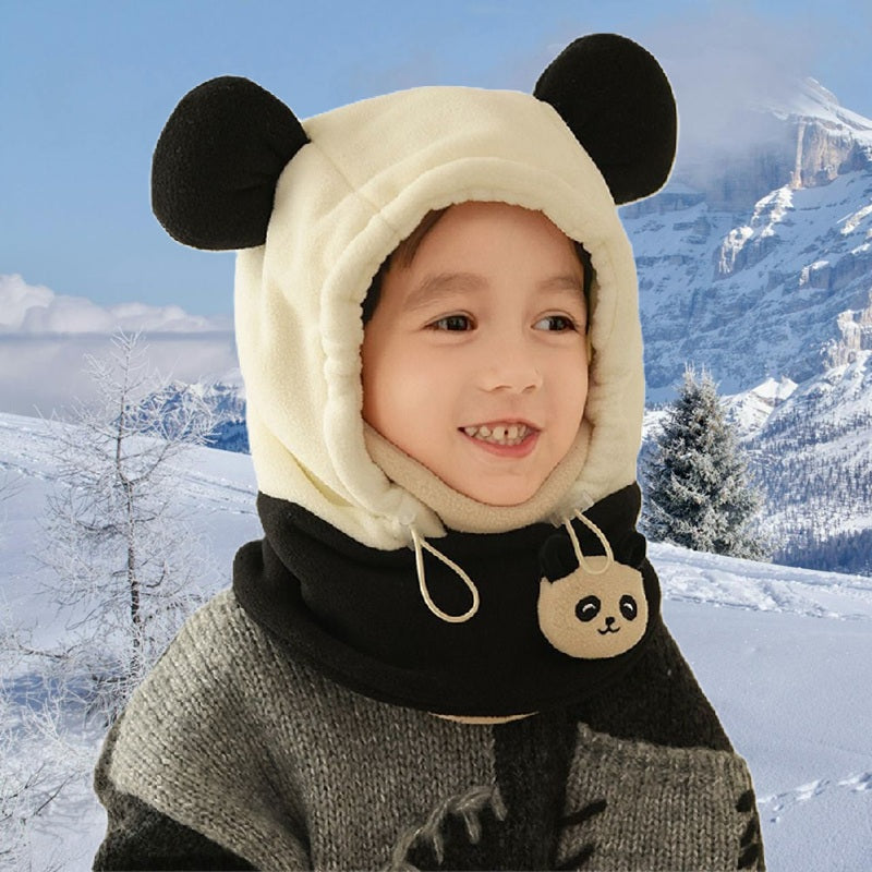 Enfant souriant habillé d'une cagoule grand froid enfant noire et blanche avec oreilles rondes, col chaud orné d'un motif panda, debout devant un décor hivernal avec montagnes et neige.