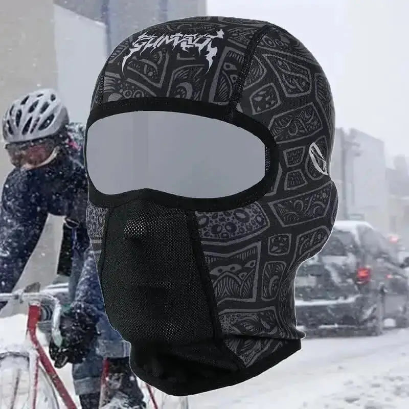 Cagoule hiver vélo noire avec motifs tribaux, conçue pour protéger les cyclistes du froid et du vent, portée par un cycliste dans un environnement urbain enneigé avec circulation et conditions hivernales difficiles.