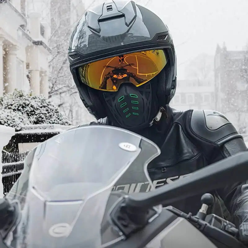Motard portant une cagoule moto hiver anti-buée avec un casque noir, des lunettes teintées jaunes et une tenue en cuir, roulant sous la neige pour une protection optimale contre le froid et la buée.