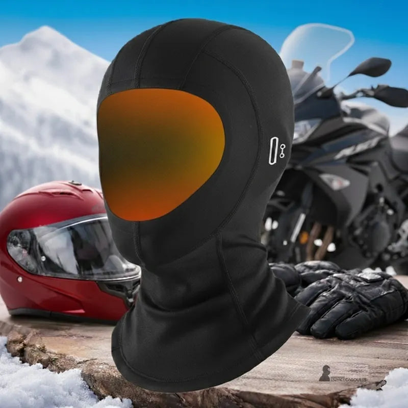 Cagoule moto hiver MaxVision posée sur une table en bois avec casque rouge, gants noirs et moto en arrière-plan enneigé, vue produit en situation extérieure - Espace Cagoules